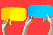 Human Hands Holding Yellow, Blue and White Speech Bubbles Over Light Orange Red Background - Fun Balloon speech bubble concept