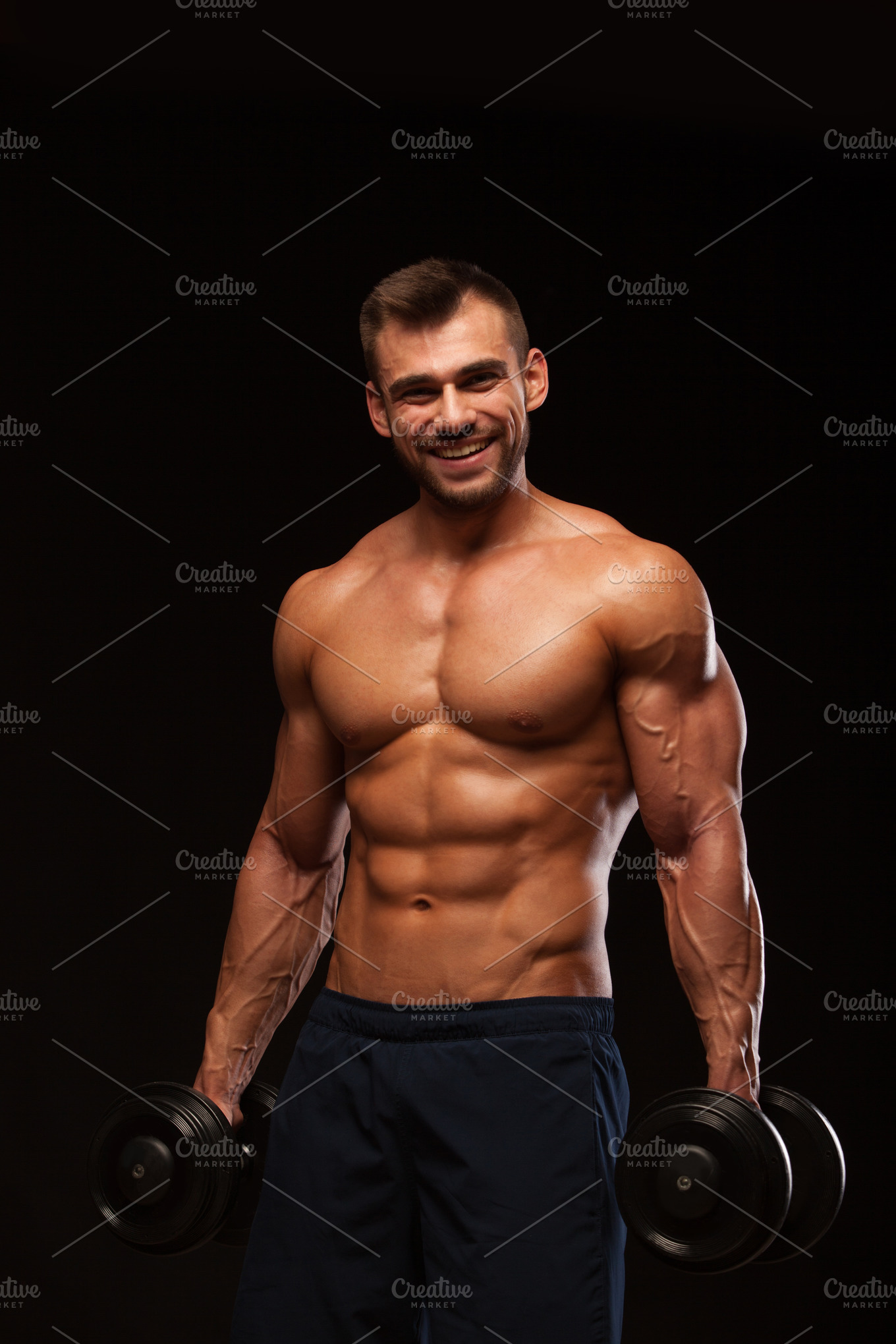 Handsome athletic man in gym is pumping up muscles with dumbbells in a ...