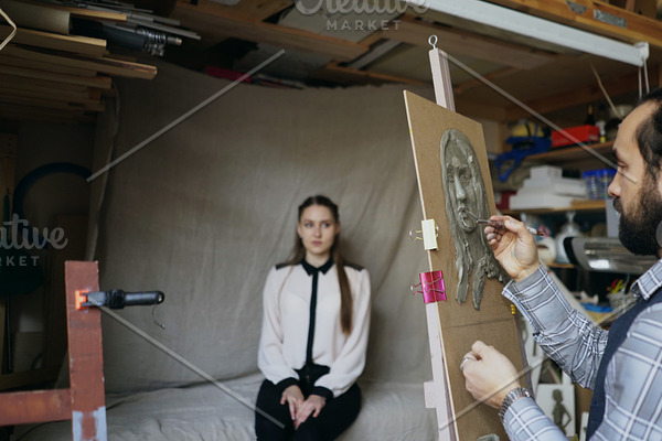 Sculptor creating sculpture of human's face on canvas while young woman posing to him in art studio