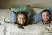 Top view of smiling couple having fun in bed hiding under blanket and looking into camera