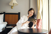 Pretty young woman in bathrobe chatting on tablet computer sitting on chair in hotel room